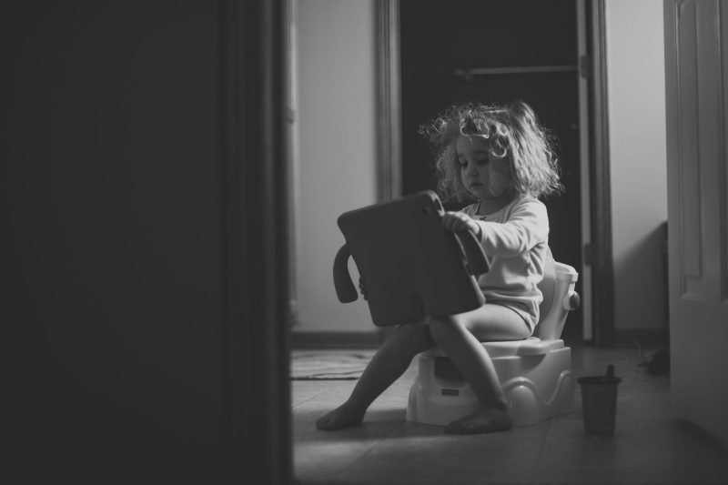 Black and white image of a girl sitting on a potty holding a toy