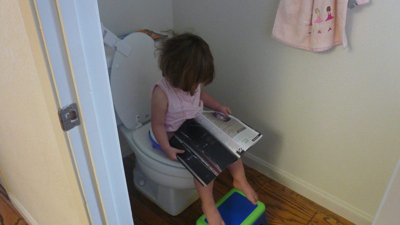 Girl sitting on a toilet with her feet on a stool reading a magazine