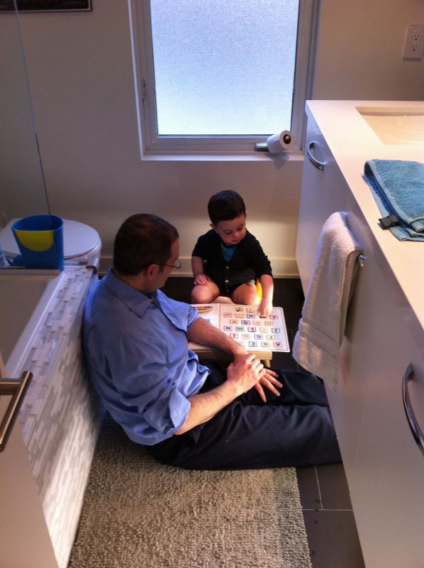 Little boy and his dad sitting on the floor in the bathroom reading a book. Boy is sittong on a potty.