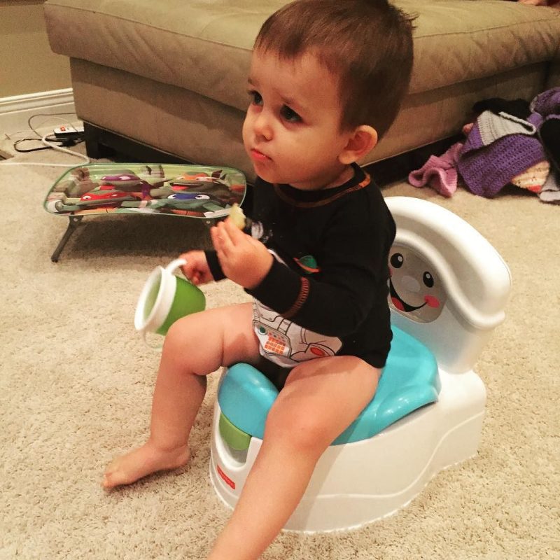 Toddler boy sitting on a potty in a lounge room holding food in one hand and a sippy cup in the other hand. Boy has a confused look on his face.