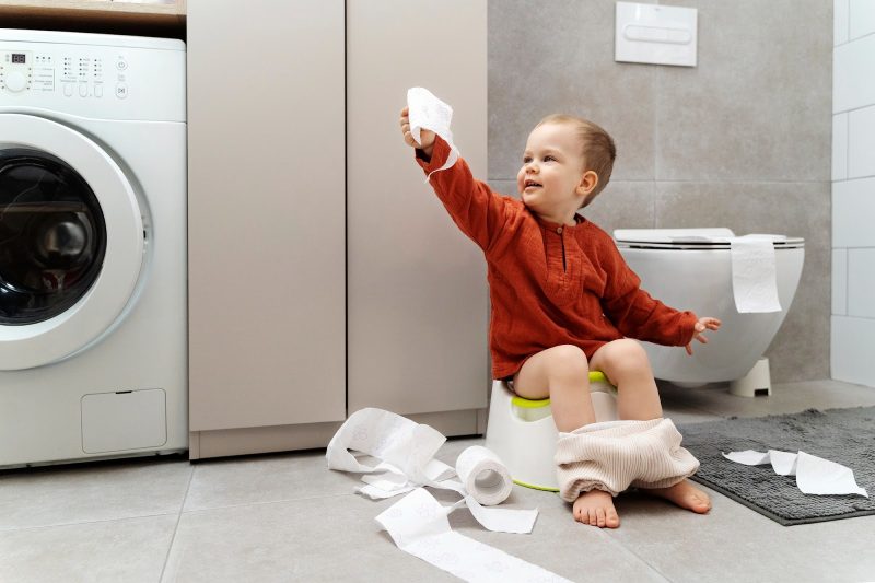 Boy sitting on a potty with toilet paper on the floor. He is holding a piece of toilet paper