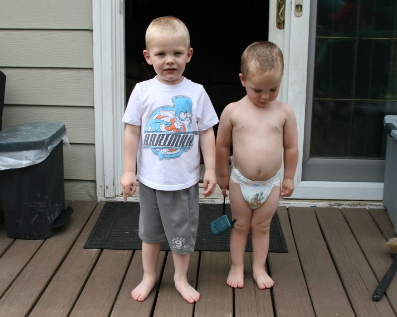 Two toddler boys standing next to each other, the younger one wearing a nappy